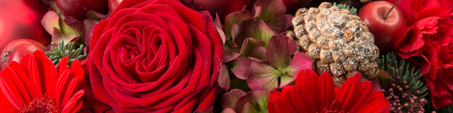 En tæt sammensætning af røde blomster, herunder roser og gerberaer, ledsaget af grønne planter og dekorationer som æbler og en fyrrekogle. Farverne skaber en festlig og varm julestemning.